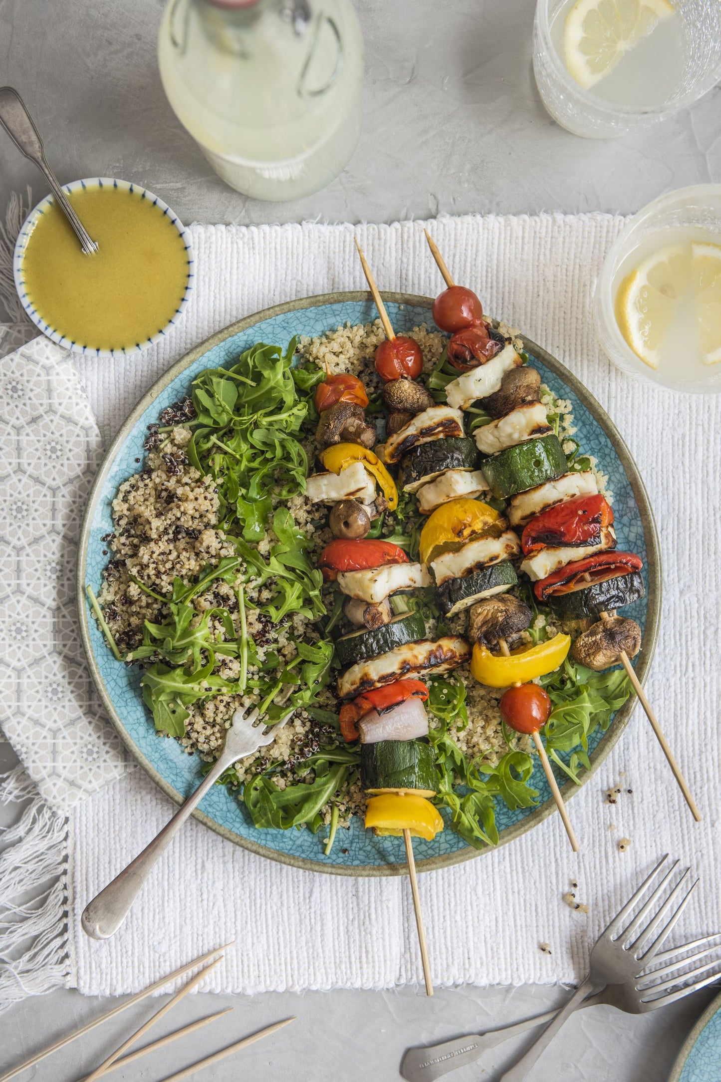 Plate of quinoa with rocket and three skewers with pepper, courgette, halloumi, tomato and mushroom. A jar of salad dressing on the side.