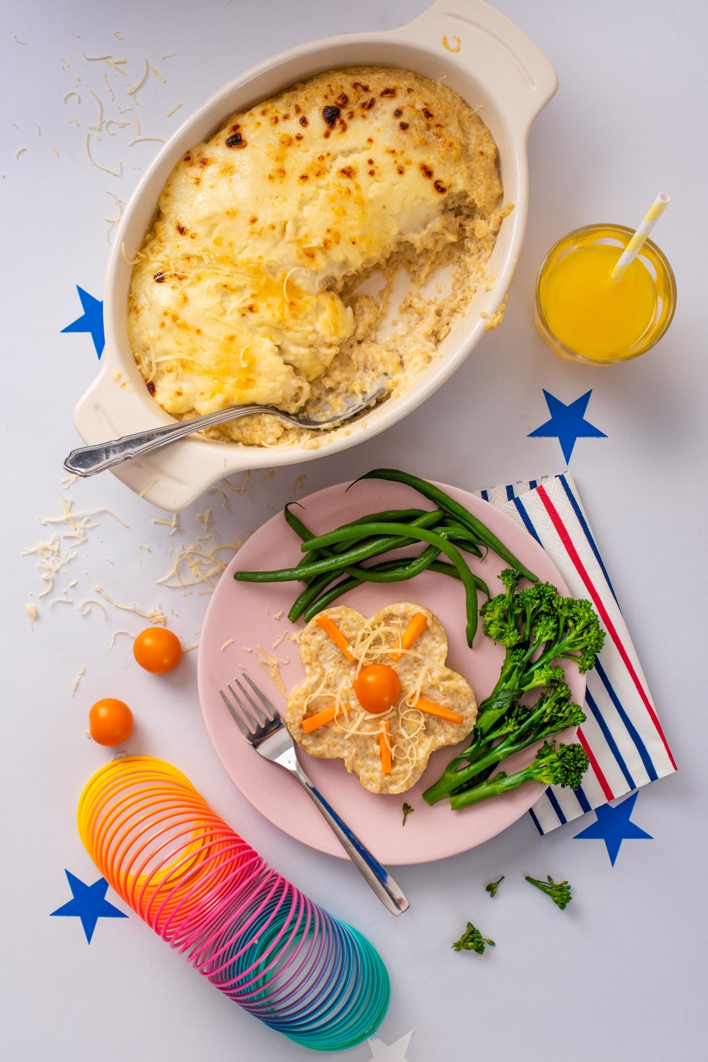 Dish of cheesy quinoa bake next to a plate of greens and a slice of cheesy quinoa. A slinky and kids toys placed around the background
