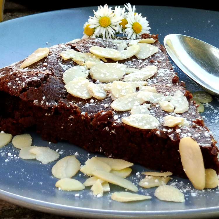 Slice of gooey quinoa chocolate cake with icing sugar and flaked almonds dusted across, daisies on the side of the plate