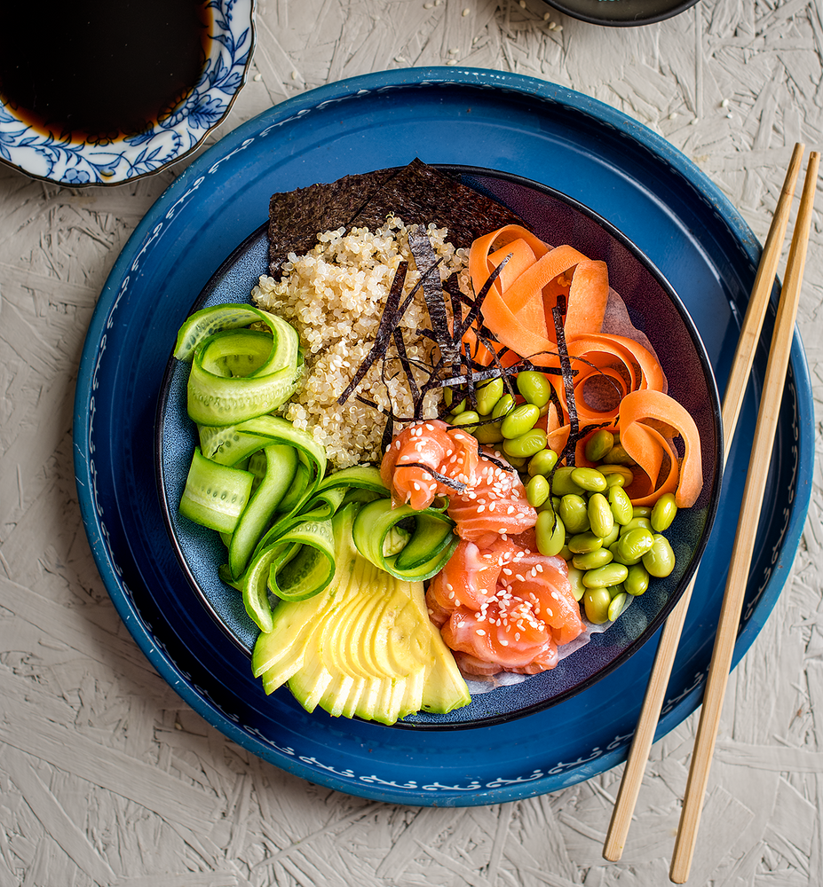 Blue bowl topped with seaweed, quinoa, avocado, cucumber, edamame beans and swirls of carrot and salmon bites 